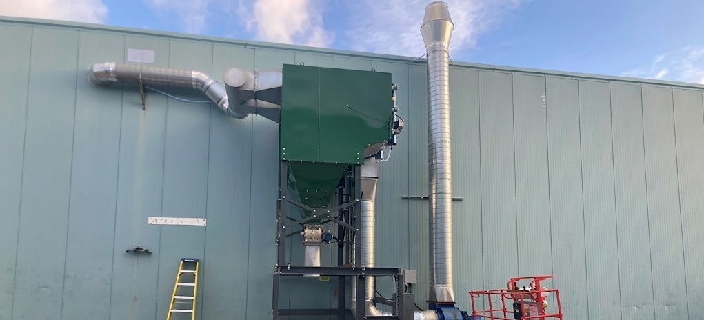 A light blue industrial building with a large green hopper, silver exhaust pipe, corrugated chimney, and a red scissor lift parked alongside.