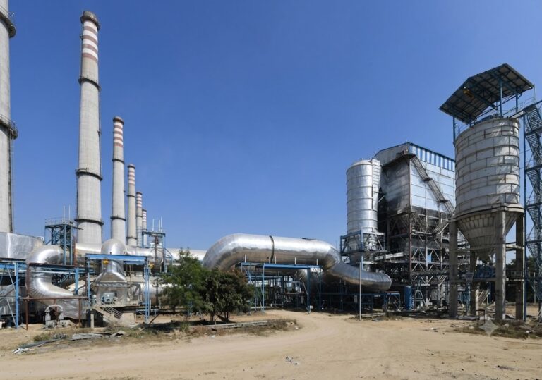A daytime wide-angle photograph of an industrial processing facility under a clear blue sky. On the left side of the frame stand several tall concrete smokestacks with distinctive red and white stripes near the top. Large, insulated silver metal pipelines snake through the center of the yard, supported by blue steel frameworks. To the right, a multi-story industrial building features external staircases, structural metal framing, and a prominent vertical cylindrical storage silo.