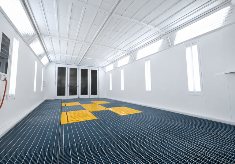 Inside an empty, bright white professional automotive spray paint booth featuring a grated floor with yellow accent panels, vertical wall lights, and large glass double doors at the back.
